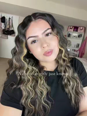 A selfie-style shot featuring a young woman. She is positioned in the center, her face is the main focus. Her wavy hair is a mix of dark and light brown tones. She has full lips with visible lipstick or gloss, and her eyebrows are well-defined. She's wearing a dark-colored shirt. The background includes a glimpse of a bedroom setting with a decorated shelf and various items. Text overlayed at the bottom reads, 'A christian girly just knows.'