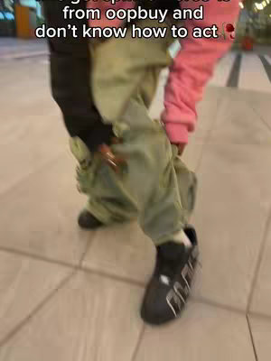 Image shows a person from the knees down, wearing a colorful outfit. The person is standing on a tiled surface, with the focus on their legs and shoes. The outfit consists of green cargo pants, a black top, and a pink long-sleeve shirt. The shoes are primarily black with white accents. Text overlayed on the upper portion of the image reads, "I got drip is from oopbuy and don't know how to act."