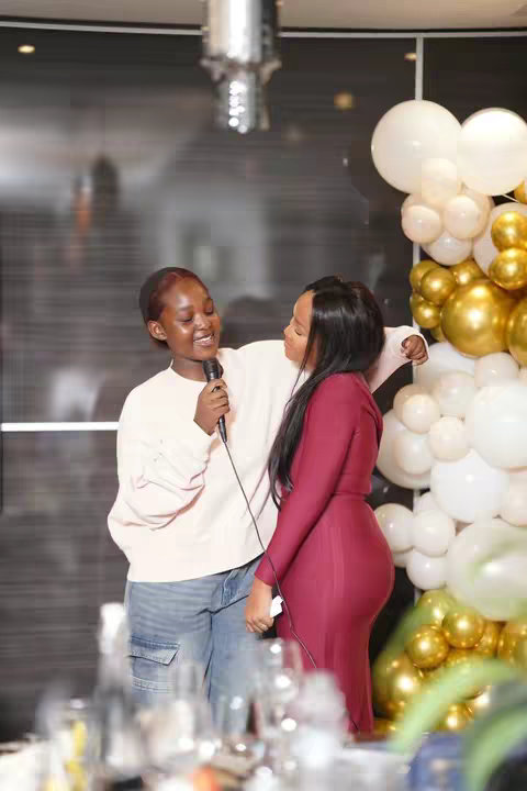 Two young women are standing close together and smiling, one holding a microphone. The woman on the left is wearing a light pink sweater and blue jeans, while the woman on the right is wearing a long, deep maroon dress. Behind them on the right are white and gold balloons. The background is blurred, suggesting an indoor event.