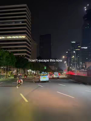 A nighttime cityscape with a road in the foreground and tall buildings on the sides. The image is taken from a driver's perspective, with streetlights illuminating the scene. The road is wet, suggesting recent rain; vehicles are visible in the distance.