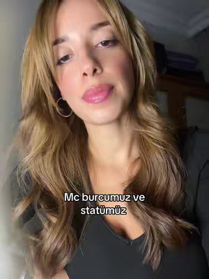 A woman with long, wavy blonde hair is the focus. She has pink lip gloss and is looking towards the camera. The text overlayed says 'Mc burcumuz ve statümüz'. The background is softly out of focus, suggesting an indoor setting. She is wearing a black top.