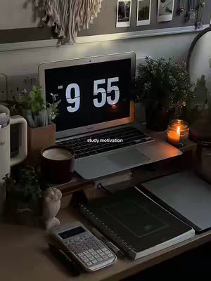 A desk setup with a laptop displaying '9:55' on the screen. The desk is cluttered but stylish, with books, a candle, plants and stationery. The image has a warm, cozy, and inspiring atmosphere, perfect for study motivation.