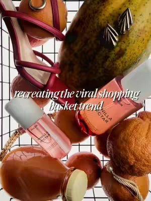 A vertical image of a shopping basket filled with various items. A red high-heeled shoe is placed prominently, with a section of a yellow fruit at the back. Several bottles of perfume, labeled "Vanilla Nectar", are scattered among brown fruits. Golden earrings and a gold chain are also present, with text overlayed at the center reads "recreating the viral shopping basket trend". The color palette is warm, featuring reds, oranges, beige, and white from the basket.