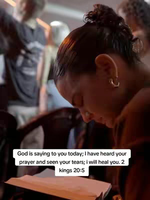 Close-up shot of a person, seen from the side, with a dark hairstyle and wearing a hoop earring. They are looking down at a book, illuminated by light. In the background, other blurred figures are present. A text overlay at the bottom showcases a quote from the Bible.