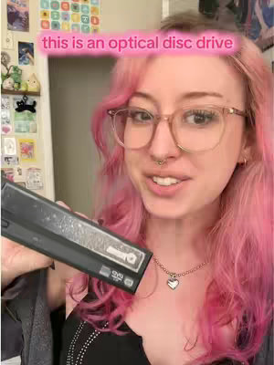 A selfie of a young woman with pink hair and glasses holding a black optical disc drive, smiling at the camera. The background is a room with various items displayed, and a pink banner with the text 'this is an optical disc drive' is overlaid on the image.