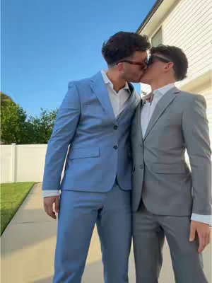 Two men in formal suits are captured in a kissing embrace, facing each other. The man on the left, wearing a light blue suit and sunglasses, has his arms around as he kisses the other man. The other man, in a grey suit and shades, mirrors the embrace. They are on a pathway with a grassy area and a white fence visible in the background, set against a blue sky.