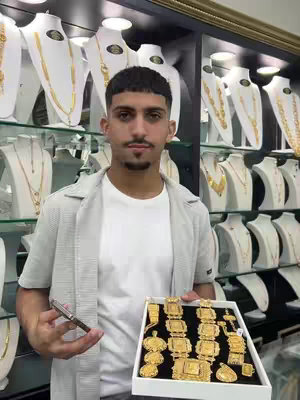 A man in a light gray shirt and white t-shirt stands in a jewelry store, holding a tray of gold jewelry. The background features shelves displaying various gold necklaces and jewelry pieces. The lighting is bright, and the layout is organized.