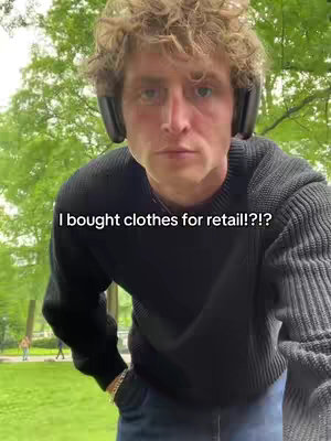 A young man with curly hair wearing a black sweater and headphones is looking at the camera from a slightly low angle. Text is overlaid at the bottom: 'I bought clothes for retail!?!?'. The background includes green foliage, suggesting an outdoor setting.