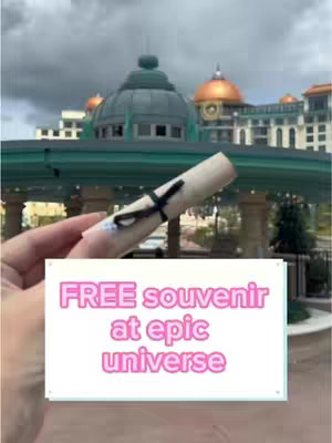 A hand holds a scroll in front of a blurred background of a building's facade and cloudy sky. A white box overlay features the text 'FREE souvenir at epic universe' in a casual pink font.
