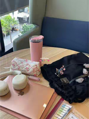 A photo of a table with several items on it, likely in a university or study setting. The items include a pink tumbler, a pencil case, a laptop, headphones, and some notebooks. The color scheme is predominantly pink with some white, black and some color from the sticky notes.