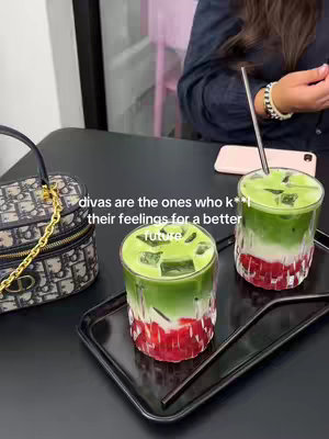 The image captures a stylish scene, possibly from a cafe or restaurant, with a black table setting. On the table, there are two glasses of layered drinks featuring a vibrant green top layer, a white middle layer, and a red bottom layer, presented on a glossy black tray alongside two straws. A designer handbag is visible, adding a note of luxury. The lighting appears soft and natural. The text overlay adds to the theme of self-care, suggesting that the image is promoting a positive mindset.