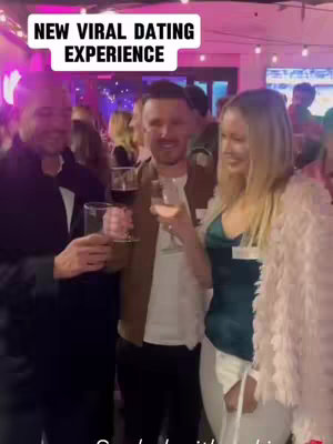 A group of three people at a bar or event, two men and a woman, are holding drinks and smiling. The background shows other people and party lights.