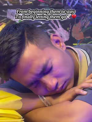A close-up shot of a man with dark hair lying down. His head is tilted slightly, and his eyes are closed. The background is soft with purple and blue hues. Text overlays are visible in the photo. His left arm is bent, with a watch bracelet on it, and he is resting his head and cheek on his folded arms. The lighting is dim, casting shadows across his face. The overall composition gives a feeling of melancholy or introspection.