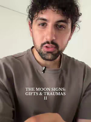 A man with dark curly hair and a beard is centered in the frame, facing the camera. He wears a gray t-shirt and a small microphone is clipped to it. The background is plain white. Text at the bottom of the frame reads: THE MOON SIGNS: GIFTS & TRAUMAS II