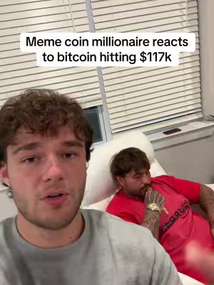 The image shows two men in a room. The man in the foreground is wearing a gray t-shirt and talking, with his head slightly turned towards the camera. Behind him, another man wearing a red shirt is seated on a white surface, engrossed in thought. The text overlay at the top reads, 'Meme coin millionaire reacts to bitcoin hitting $117k'. Behind them is a window covered with blinds.
