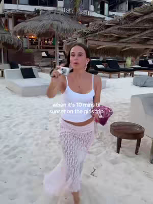 A woman walking towards the camera on a sandy beach. She is holding a camera and purse. Background features beach umbrellas, lounge chairs, and buildings.