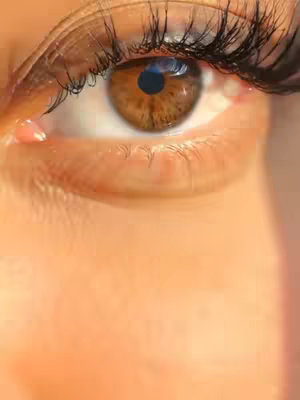 Close-up of a brown eye with long lashes. The eye is the focal point, filling most of the frame. The iris is a rich brown color with detailed texture, and the pupil is a dark circle. The eyelashes are long and dark, framing the eye. The skin around the eye is a light peach color, with slight shadows indicating the contours of the face. The overall lighting appears soft and natural, with a slight glow on the skin, and the background is blurred out.