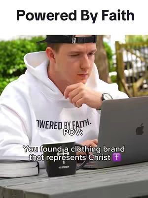 A man sits at a table outside, working on a laptop. He is wearing a white hoodie and a black cap. The image is well-lit and the background is slightly blurred, with greenery visible.
