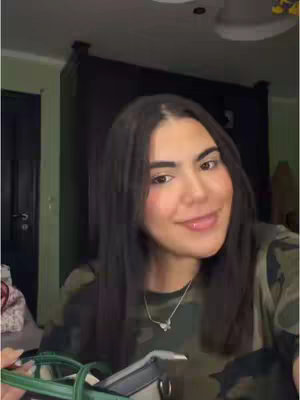 A person is shown in a selfie-style shot, smiling slightly. The person has long dark hair and is wearing a green camouflage shirt and a silver necklace with a small pendant. In their hands, a green purse with a black top is held forward. The background includes hints of a cluttered room.