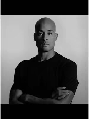 A black and white portrait of David Goggins. He is centered in the frame, with his arms crossed in front of him at chest level. He is wearing a black t-shirt. The background is a plain, light gray. The photo has high contrast and a serious tone.