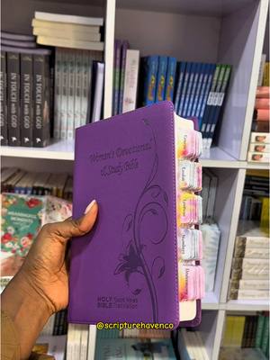 A hand holding a purple bible with tabs, in front of a bookshelf. The bible's cover has the words 'Women's Devotional & Study Bible'. The background is a bookshelf filled with books.