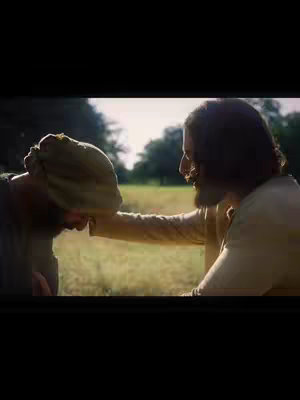 The image shows two men in a natural setting. One man, wearing a neutral-colored garment, is touching the other's head. The background suggests a grassy area with sunlight.