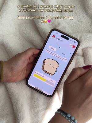 A person's hands hold a phone displaying a budgeting app interface. The app's screen features a cute cartoon of a piece of toast, with a budget progress bar. The background is a soft, white blanket. The person has dark nail polish and wears a bracelet.