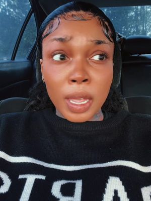 A close-up shot of a woman inside a car, looking to the side with a surprised expression. She has dark skin, slicked-back hair with baby hairs, and is wearing a black sweater. The interior of the car, including the seat and door, are visible in the background.