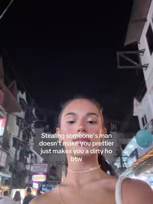 A selfie-style shot taken at night. The shot is framed with a woman's face in the foreground. The background includes buildings with various lights, suggesting a city or town environment. The sky is dark, contributing to the night atmosphere.
