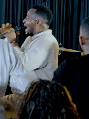 The image shows a man singing into a microphone with an open mouth, likely engaged in an enthusiastic performance. He's wearing a light-colored shirt and appears to be in a church or performance setting, suggested by the dark background and lighting. Other people can be seen in the image and at least one other person standing near the main subject. The focus is on the performer's expression and the act of singing.