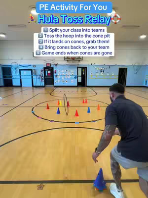 The image shows a PE teacher in a gym setting demonstrating a hula hoop toss relay game. The image is brightly lit with a wooden floor. The teacher is wearing a dark shirt, shorts, and has tattoos on his arm