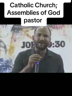 A selfie-style video featuring a man speaking with a microphone. Behind him is a poster with blurred text and the words 'JOHN 9:30'. A text overlay at the top reads 'Catholic Church; Assemblies of God pastor'. The background suggests a religious setting.
