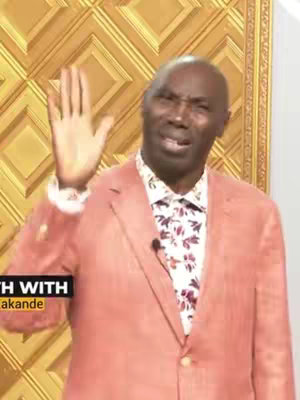 A man wearing a pink blazer and a floral shirt raises his right hand with his palm facing out. The background is a gold, ornate, geometric pattern. The image is well-lit, and the man seems to be addressing the viewer. A graphic in the lower left is partially visible with the text 'ITH WITH' and 'Akande'.