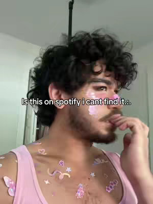A selfie-style shot featuring a young man with dark curly hair, a light complexion, and facial hair, particularly a mustache and stubble. He's wearing a light pink tank top. Decorations, including pink and white stickers in various shapes like stars, butterflies, and flowers, are adhered to his chest and shoulders.  Above the man's head, there is a black pole or stand. The background is lightly colored. The text overlay reads, "Is this on spotify I cant find it.."