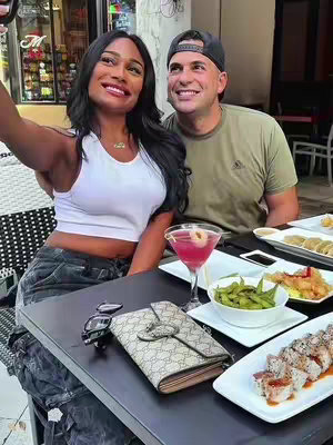 A selfie taken by a woman with a man in the background, sitting at a table with sushi, cocktails, and edamame.
