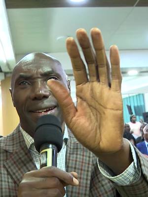 A man with a bald head and a plaid blazer is holding a microphone with one hand and waving with the other. People are in the background.