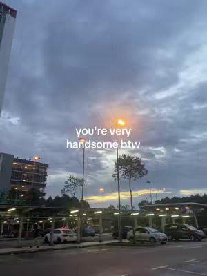 The image shows a parking lot at dusk with a cloudy sky above. The sky and the environment colors are various shades of grey and blue, with a warm glow from streetlights and the lights of a building in the distance. The text overlay is in white and centered on the image.