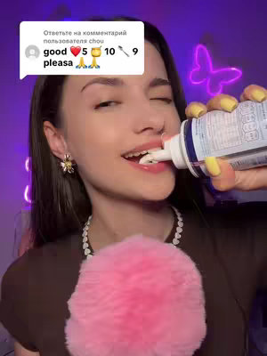 A young woman is shown from the shoulders up, holding a tube to her mouth and squeezing out a white substance. Her mouth is open, and she appears to be enjoying the product. She has dark hair, styled with a slight wave, and is wearing a brown top with a pearl necklace and flower earrings. Her nails are painted yellow. Behind her is a blurred purple background with a neon butterfly.