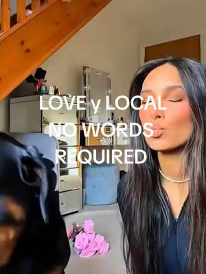 A woman with long dark hair is puckering her lips. Overlayed text reads 'Love y Local, No Words Required'. A dog's head and shoulders are visible to the left, while a partially visible room with a mirror is in the background.