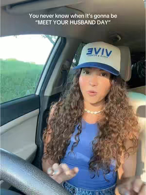 A woman with long, curly hair is seated in the driver's seat of a car, looking towards the camera. She wears a baseball cap and blue top and a necklace. The car's interior and the outdoor scenery are visible in the background. Overlay text across the top provides a message.
