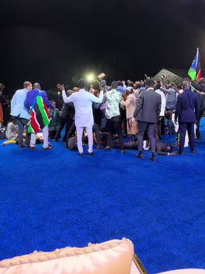 A wide shot of a large crowd gathered in a large venue, likely outdoors, with a vibrant blue floor covering most of the surface. Several people are standing or kneeling, some with their arms raised. One individual, positioned in the foreground, has both arms raised and a person next to them is holding what looks like a Kenyan flag. The background is dark, suggesting it's nighttime, with some light sources visible at the top of the frame and behind individuals