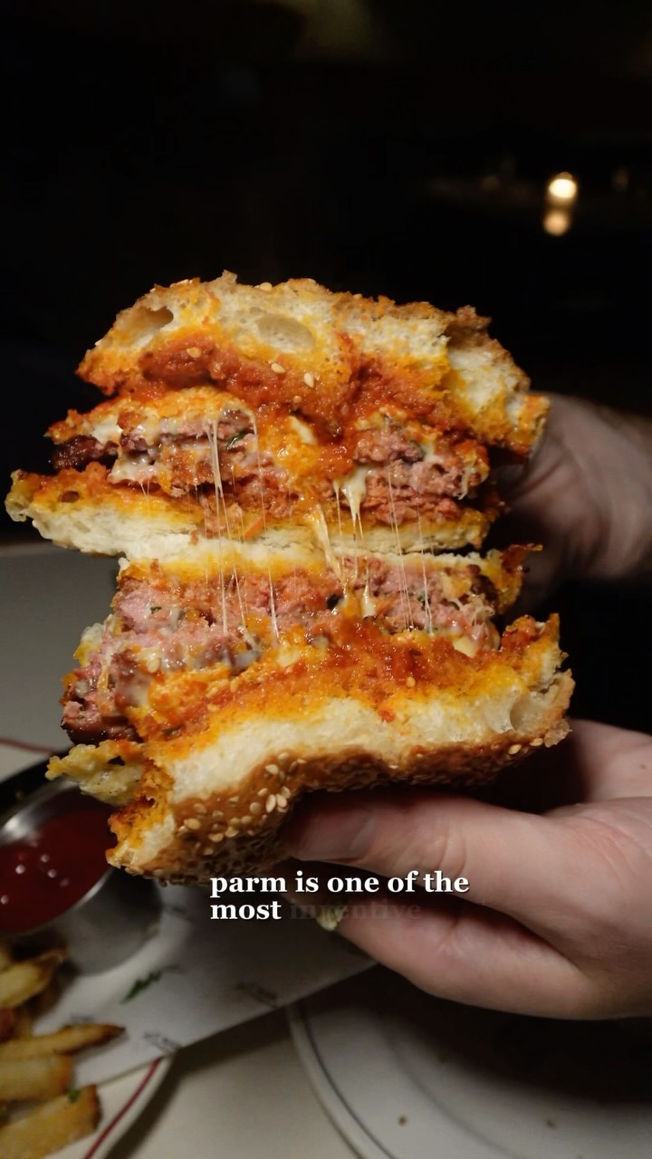 The cover image shows a close-up of a meatball parm sandwich, cut in half to reveal the filling. The sandwich has a sesame seed bun with a melted cheese and red sauce filling in the middle. A hand is holding the sandwich, placed against a dark background with some indication of other food elements like fries and ketchup.
