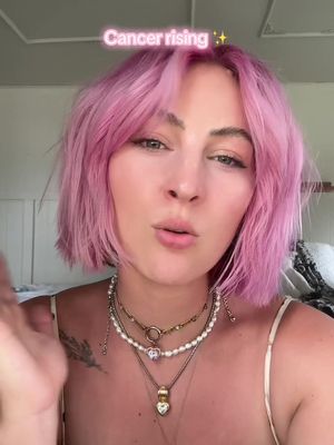 A woman with pink hair and multiple necklaces is centered in the frame. Over her head, the text "Cancer rising" is visible. The woman is looking towards the camera, and the background appears to be an interior setting possibly a bedroom.