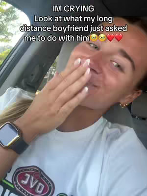 A young woman in a car, partially covering her mouth with a hand. Text overlays read "IM CRYING" and "Look at what my long distance boyfriend just asked me to do with him" with heart and crying face emojis. She appears surprised and emotional. The background shows a window and the interior of the car.