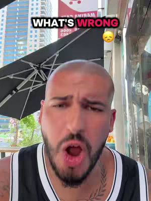 A man with a shaved head and a goatee is the focal point. He is wearing a black and white sleeveless shirt and has a tattoo on his chest. He is looking at the camera with his mouth open in an expression of surprise or shock. Above him, a graphic displays the words "WHAT'S WRONG" in a white, bold font. Next to the text, there is a sad, emoji-like face. The background appears to be an outdoor setting with an umbrella and some buildings.