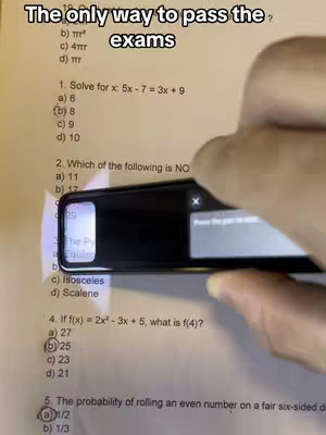 A person's hand holding a smartphone is partially visible, positioned over a piece of paper filled with mathematical questions. The phone is angled to capture a portion of the paper, with the camera lens facing downwards towards the questions.