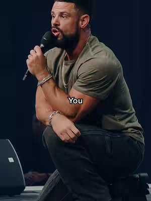 A man with short, dark hair and a beard is seated on a stage, holding a microphone. The man is wearing a green t-shirt and dark pants, with a silver bracelet. The word "You" is overlaid on the image.