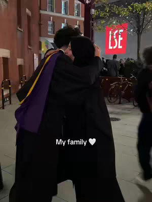 A photo captures two individuals embracing, likely outdoors given the presence of streetlights and building facades in the background. One person, on the right, has dark hair partially obscured by a black hijab. The other person is partially visible, seen from the back; they wear a graduation gown with a purple and gold sash. The words 'My family' are written in white text with a heart symbol.