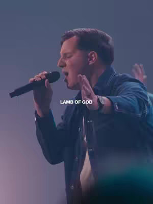 The cover image depicts a man singing into a microphone with his eyes closed and his hands at the height of his shoulders. The image is bathed in a cool, purple-blue wash. Text overlayed in white across the center reads "LAMB OF GOD".