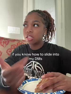 A young woman is shown from the chest up. She is speaking, and the image has text overlaid which reads, "if you know how to slide into my DMs". The background shows the inside of a room with a small portion of decorated sofa and white doors.